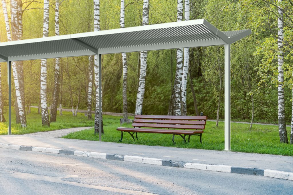 Sun shade structure protecting bench in community park setting, at the edge of a parking lot.