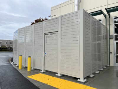 A gray solid equipment screening system with a built-in gate and two yellow bollards in a building's parking lot.