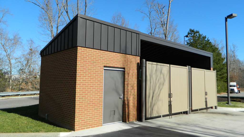 Modern public restroom building with brick exterior, black metal standing seam roof, and beige composite panel privacy screens with metal frame concealing outdoor shower or utility area