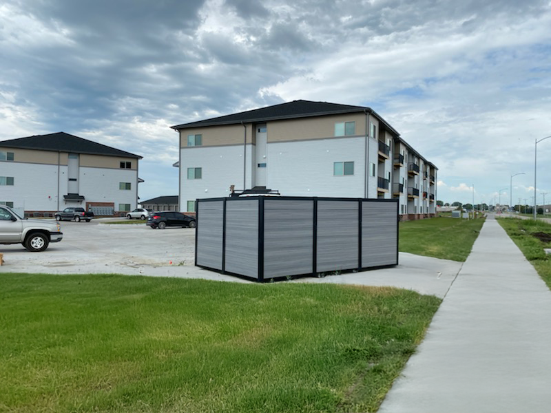 Modern composite dumpster enclosure with horizontal slat panels in gray and black aluminum frame concealing waste receptacles at multi-family residential apartment complex with lawn landscaping