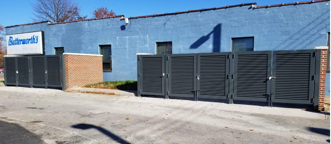 Commercial aluminum louver fence panels and gates installed along blue block building exterior with Butterworth's business signage. Industrial louvered privacy fencing system featuring horizontal slat design, multiple access gates, and professional installation on concrete pad. Modern commercial fence screening with aluminum louver construction, brick accent columns, and secure gate hardware for business property perimeter security and aesthetic enhancement.