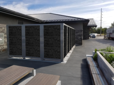 Modern commercial building exterior featuring decorative stone-textured architectural screens and mechanical equipment screens with white aluminum frames, creating a cohesive perimeter enclosure along concrete walkway with integrated landscaping planters
