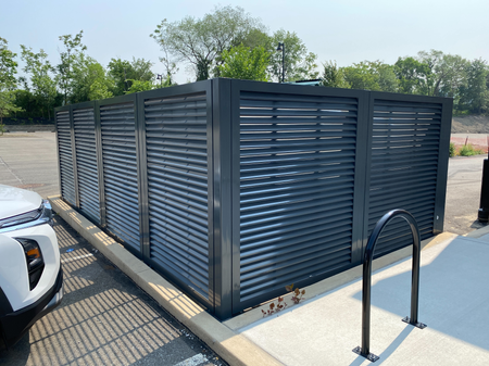 Modern commercial dumpster enclosure with dark gray horizontal louvered aluminum panels and hinged access gates concealing waste receptacles on concrete pad with black safety handrails